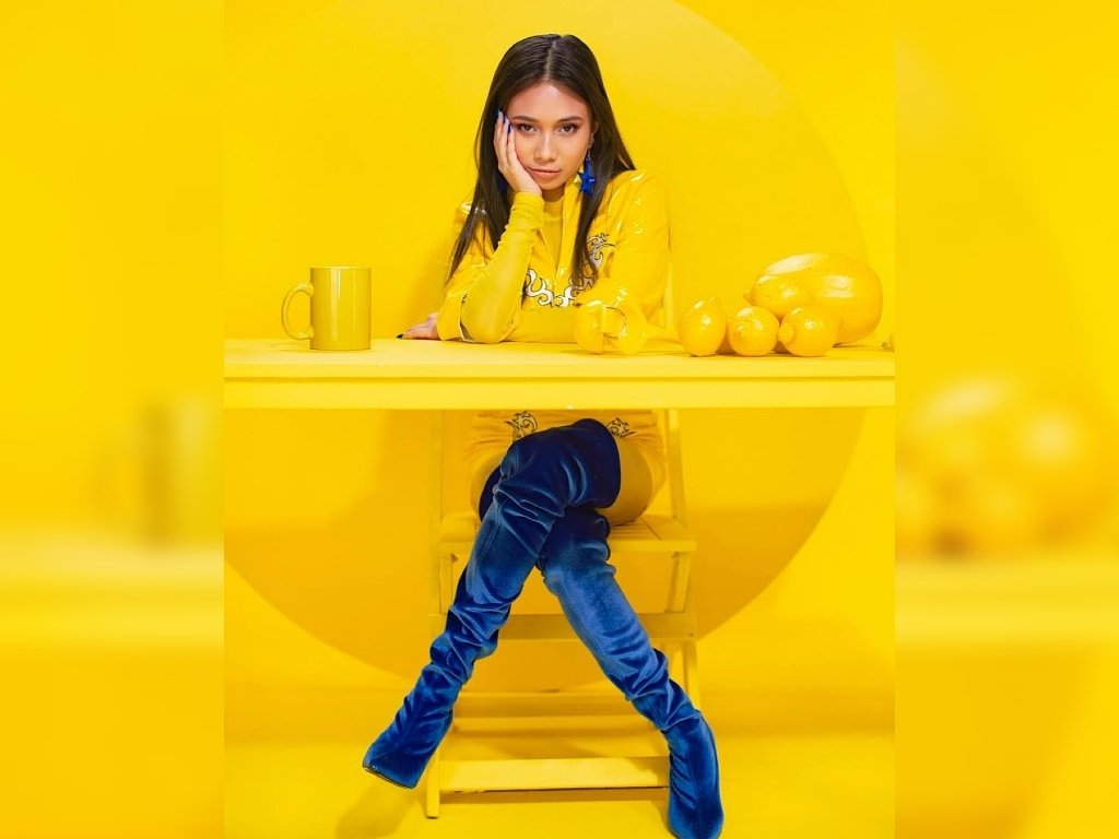 Niki mengenakan pakaian berwarna kuning serta boots warna biru sedang duduk dengan salah satu tangannya di taruh di pipi